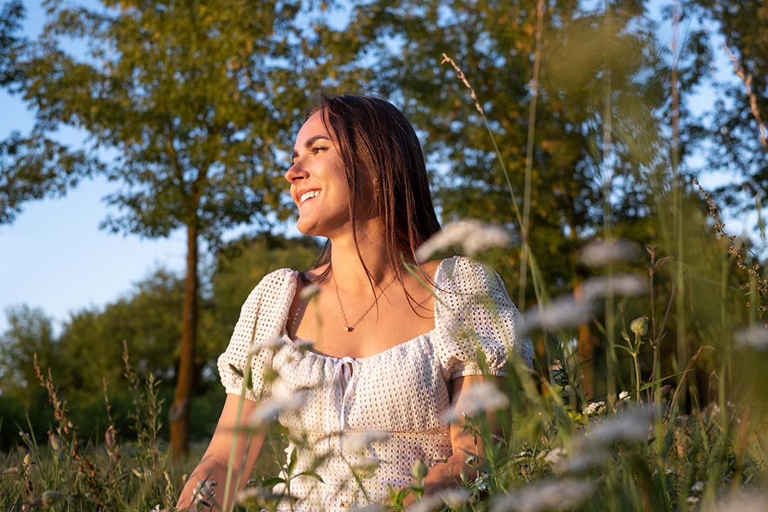 Imagen de una mujer sonriendo, sentada en el pasto, usando un vestido blanco con hombreras, al atardecer, en el campo