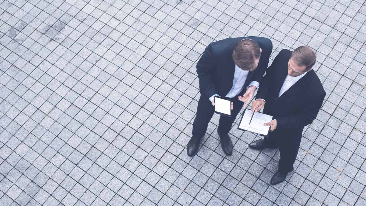 Imagen de dos ejecutivos conversando y viendo sus apuntes, en una vista aérea en un patio con baldosas de cemento