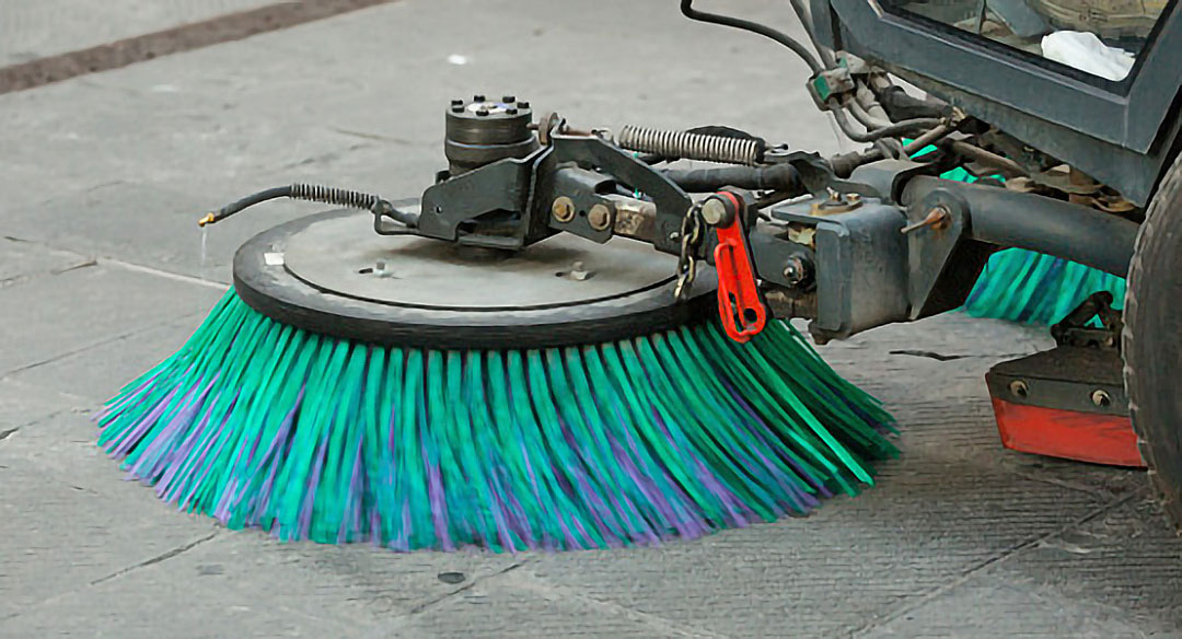 Imagen de una barredora de calles con enfoque en las cerdas limpiadoras, de color turquesa y verde, en una calle