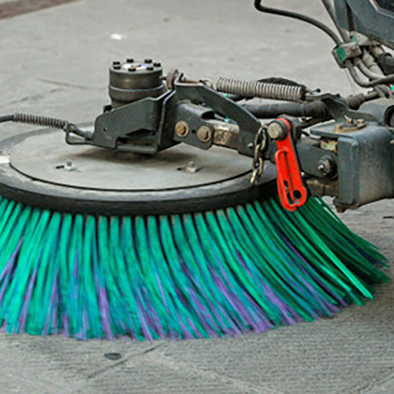Imagen de una barredora de calles con enfoque en las cerdas limpiadoras, de color turquesa y verde, en una calle