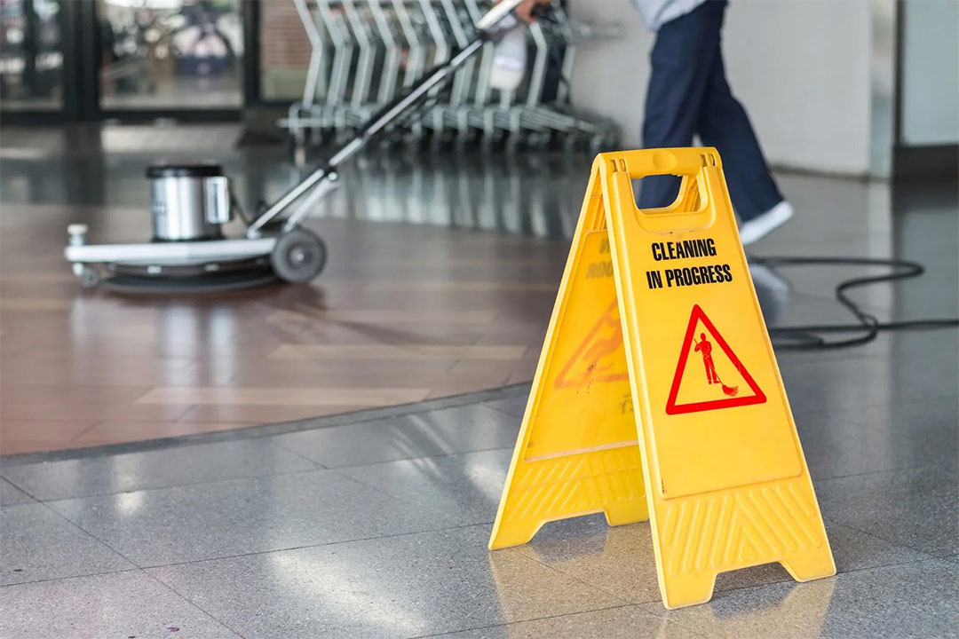 Imagen de un letrero triangular con texto de advertencia por realización de limpieza y una máquina limpiadora de pisos siendo utilizada por una persona